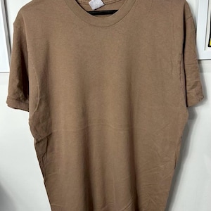Könnte beinhalten: Ein kurzärmeliges, braunes T-Shirt mit Rundhalsausschnitt. Das T-Shirt hängt an einem schwarzen Kleiderbügel. Der Stoff ist einfarbig. Ein einfacher, lässiger Oberteil-Stil.