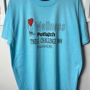 May include: Pale blue t-shirt with the text "Wellness Potlatch Fitness Challenge 1994 Brainerd, MN". The shirt features a graphic of a heart with legs. The shirt has a crew neck and short sleeves.