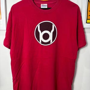 May include: A red t-shirt featuring a white and maroon circular graphic. The graphic is a stylized symbol. The t-shirt is hanging on a black hanger against a white background. The shirt is made of a soft material.