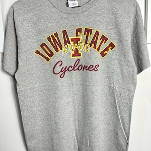 Può includere: Maglietta grigia con un logo Iowa State Cyclones in marrone e giallo. Il logo è a forma di arco con le parole "Iowa State" in giallo e "Cyclones" in marrone.