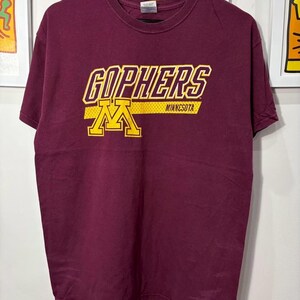 May include: Maroon t-shirt with the word "GOPHERS" in gold and yellow block letters. Below the word "GOPHERS" is a gold and yellow "M" logo with the word "MINNESOTA" in gold letters.