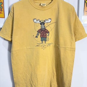 May include: A yellow t-shirt with a cartoon moose wearing sunglasses and a fishing hat. The moose is holding a fishing rod and a fish. The text "All caught up in Vermont" is printed below the moose.