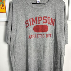 Op de afbeelding: Grijs T-shirt met een rode grafische print met de tekst "SIMPSON ATHLETIC DEPT".