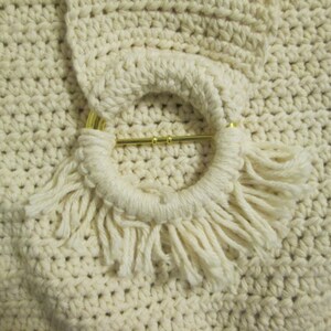 May include: Close-up of a cream-colored crocheted bag with a circular handle. The handle is adorned with a gold-colored metal bar and fringe. The bag's texture is visible, showcasing the detailed crochet work.