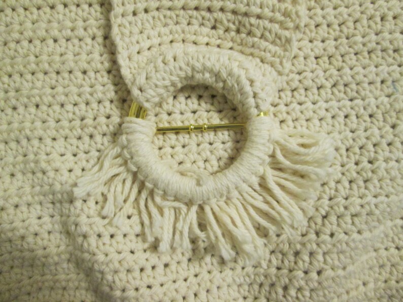 May include: Close-up of a cream-colored crocheted bag with a circular handle. The handle is adorned with a gold-colored metal bar and fringe. The bag's texture is visible, showcasing the detailed crochet work.