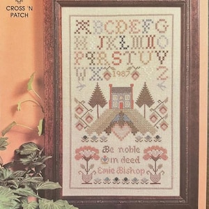 May include: A framed cross-stitch sampler titled Noble Deed by Cross 'N Patch. The design features an alphabet, the year 1987, a house, trees, and floral motifs in muted tones. The text reads Be noble in deed and the name Emie Bishop.
