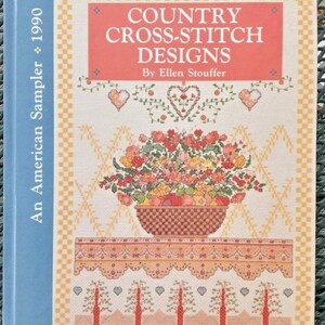 May include: A book titled "Country Cross-Stitch Designs" by Ellen Stouffer. The cover features a cross-stitch design of a basket overflowing with flowers and fruit, with a decorative border and the text "An American Sampler 1990".