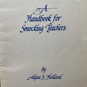 May include: A white book cover with blue text that reads "A Handbook for Smocking Teachers by Allyne S. Holland".