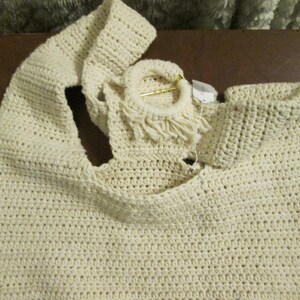 May include: Cream-colored, handmade crochet tote bag. The bag features a large, open design with a wide strap and a circular handle with fringe. The crochet stitch creates a textured surface. A small tag is attached to the handle.