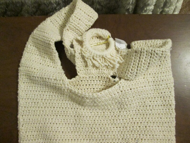 May include: Cream-colored, handmade crochet tote bag. The bag features a large, open design with a wide strap and a circular handle with fringe. The crochet stitch creates a textured surface. A small tag is attached to the handle.