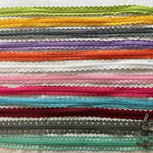 May include: A collection of colorful lace-edged zippers in various shades, including green, yellow, gray, purple, orange, white, pink, red, blue, and teal. The zippers are arranged horizontally, showcasing their decorative scalloped edges and metal pulls.