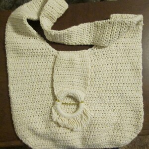 May include: Cream-colored crochet shoulder bag with a wide strap. The bag features a circular decorative element with fringe and a gold-colored ring. The bag is made of a thick, textured yarn.