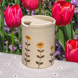May include: A cylindrical ceramic container with a speckled off-white finish, featuring a hand-painted design of three sunflowers with brown stems and green leaves. The container has a notched rim and a flat lid, positioned upright.