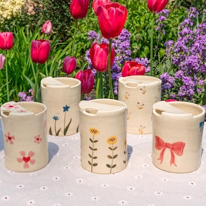 May include: Five off-white ceramic travel mugs with lids, each decorated with a different floral design. The cylindrical mugs are displayed on a white tablecloth with a floral pattern. The background features vibrant red tulips and other colourful flowers.
