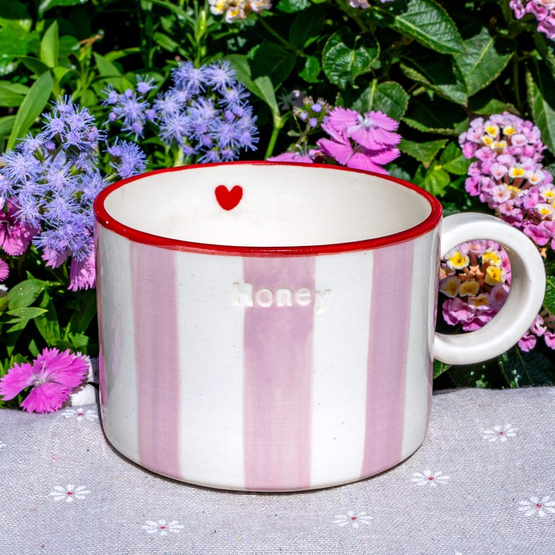 May include: A striped ceramic mug with pink and white vertical stripes. The rim is red, and a small red heart is inside the rim. The word "Honey" is embossed on the side. The mug has a white handle.