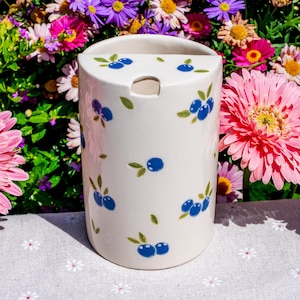 May include: A white ceramic utensil holder with a cylindrical shape. The holder is decorated with blue blueberry clusters and green leaves. The background features a variety of colorful flowers, including pink gerbera daisies and purple asters.