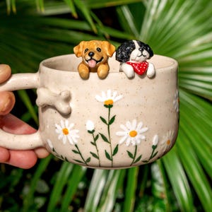 Puede incluir: Una taza de cer&aacute;mica color crema moteada con margaritas blancas pintadas a mano con centros amarillos. Dos peque&ntilde;as figuras de perros esculpidas, una dorada y otra blanca y negra con un pa&ntilde;uelo rojo de lunares, asoman por el borde. Asa en forma de hueso.