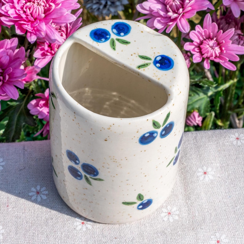 May include: A speckled ceramic holder with a curved top and a diagonal opening. The holder is decorated with blue blueberry clusters and green leaves. The background features pink flowers.
