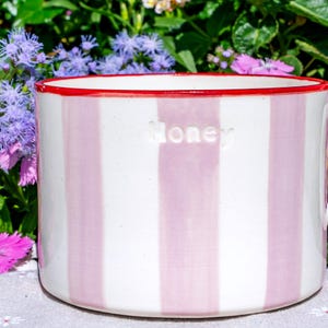 May include: A striped ceramic honey pot with pink and white vertical stripes. The rim is red, and the word "Honey" is embossed on the front. The pot is cylindrical and sits on a white surface, with flowers in the background.