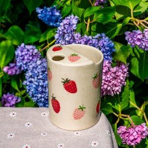 Può includere: Un portaspazzolino in ceramica color crema con un motivo a fragole. Il supporto ha una forma cilindrica con una parte superiore divisa e un foro per uno spazzolino. Le fragole sono rosse e rosa, con foglie verdi. Lo sfondo presenta fiori viola e blu.