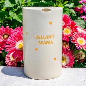 May include: A cream-colored ceramic container with the text "KELLAN'S MAMA" in gold lettering, adorned with small gold heart accents. The container is cylindrical with a small opening at the top. Bright pink gerbera daisies with yellow centers are in the background.