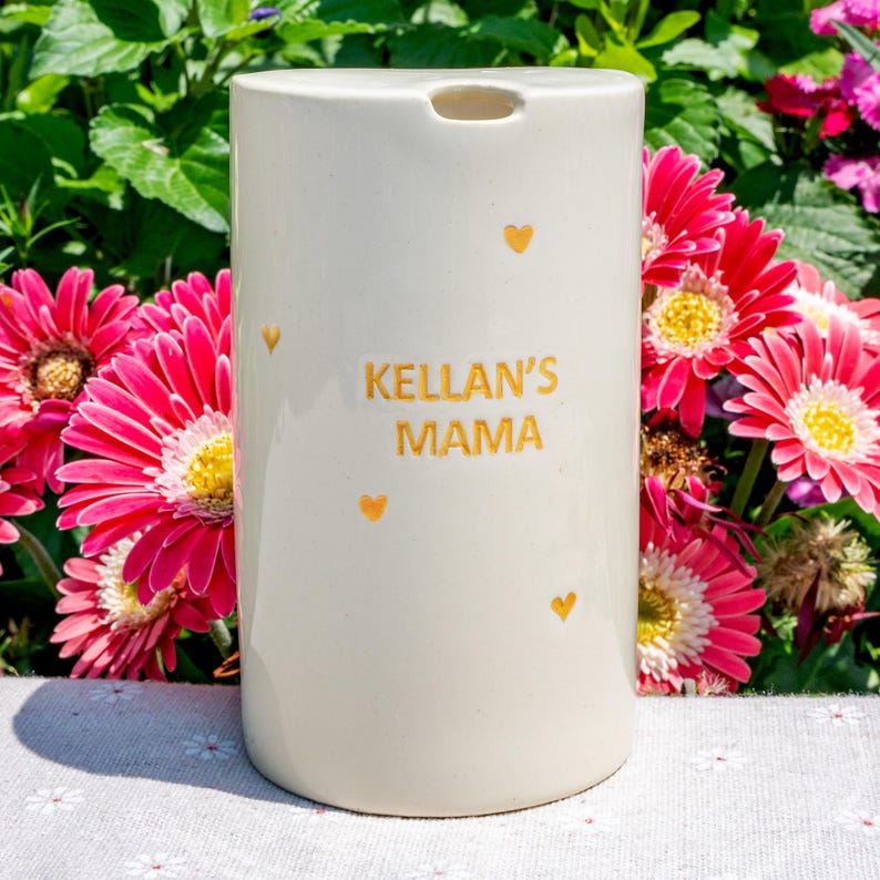 May include: A cream-colored ceramic container with the text "KELLAN'S MAMA" in gold lettering, adorned with small gold heart accents. The container is cylindrical with a small opening at the top. Bright pink gerbera daisies with yellow centers are in the background.
