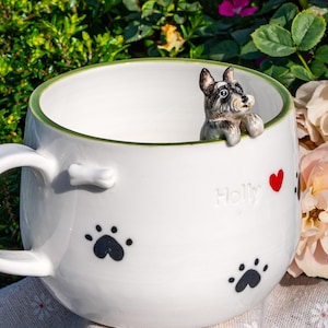 May include: A white ceramic mug with a green rim, featuring a three-dimensional miniature Schnauzer dog figurine peering over the edge. The mug is decorated with black paw prints, a small white bone near the handle, a red heart, and the name Holly.