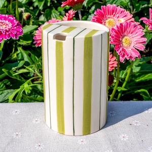 May include: A cylindrical ceramic utensil holder featuring a cream-colored base with vertical lime green stripes and thin dark green pinstripes. The top edge has a semi-circular cutout, and the surface has a smooth, glossy finish throughout.