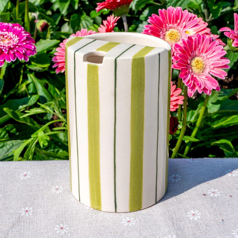 May include: A cylindrical ceramic utensil holder featuring a cream-colored base with vertical lime green stripes and thin dark green pinstripes. The top edge has a semi-circular cutout, and the surface has a smooth, glossy finish throughout.