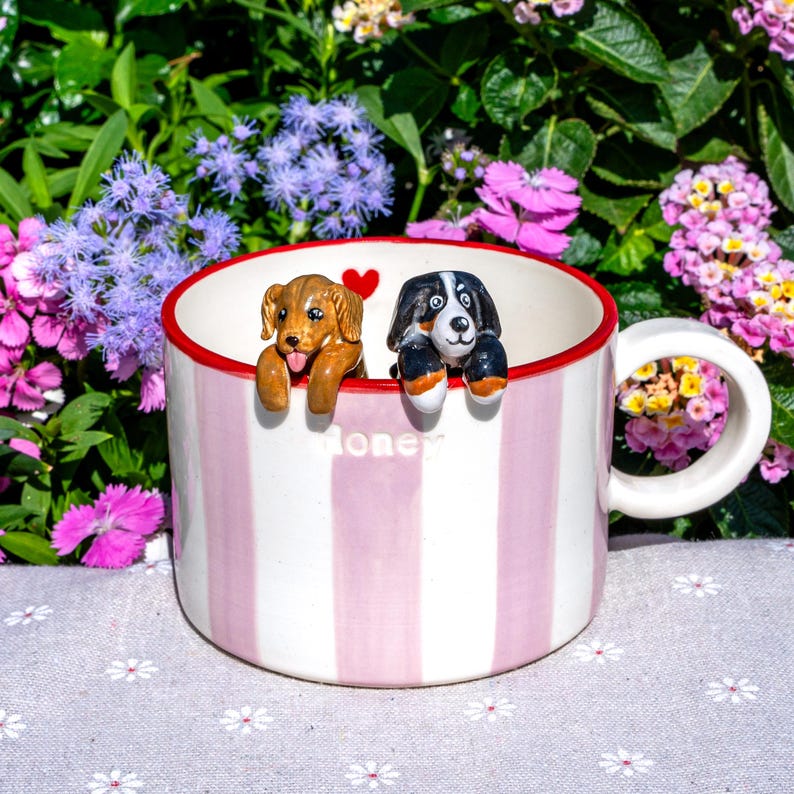 May include: A striped ceramic mug in white and pink, with a red rim. Two dog figurines, one brown and one black and white, are attached to the rim. The word "Honey" is printed on the mug. The background features flowers.
