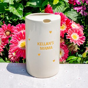 May include: A cream-colored ceramic tumbler with the text "KELLAN'S MAMA" in gold lettering, along with small gold heart accents. The tumbler has a rounded top with a small opening and is set against a backdrop of pink flowers and green foliage.