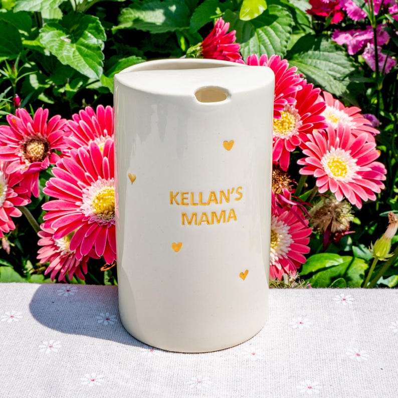 May include: A cream-colored ceramic tumbler with the text "KELLAN'S MAMA" in gold lettering, along with small gold heart accents. The tumbler has a rounded top with a small opening and is set against a backdrop of pink flowers and green foliage.