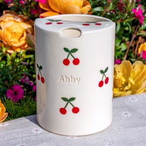 May include: A cylindrical white ceramic utensil holder featuring a repeating pattern of small red cherries with green stems and leaves. The name Abby is embossed in the center of the front surface. The top edge has a small cutout notch.