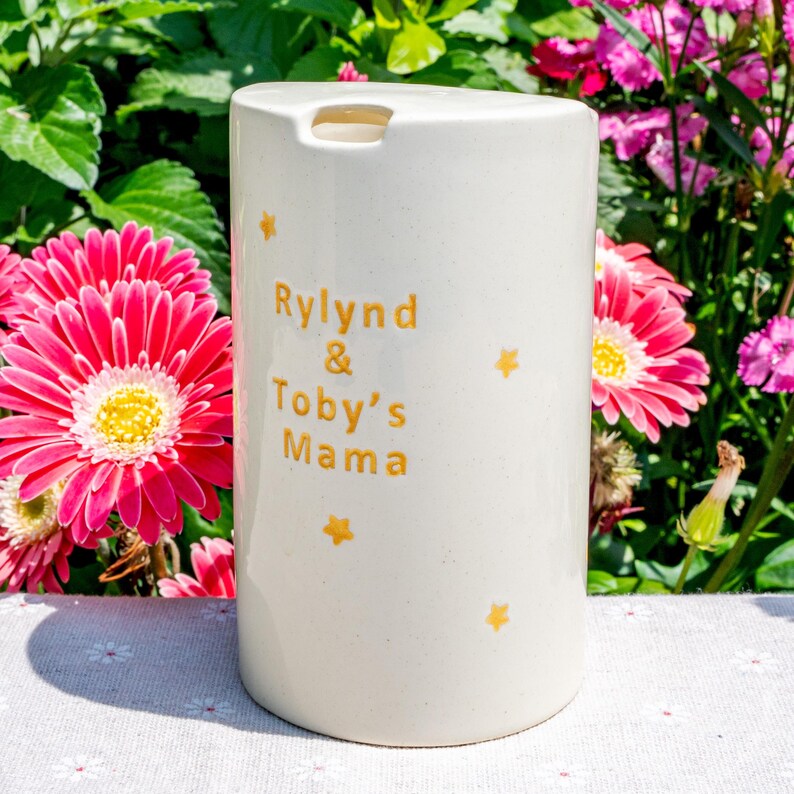 May include: Cream-colored ceramic tumbler with the text "Rylnd & Toby's Mama" in gold lettering, along with gold star accents. The tumbler has a rectangular opening at the top. The background features pink flowers and green foliage.
