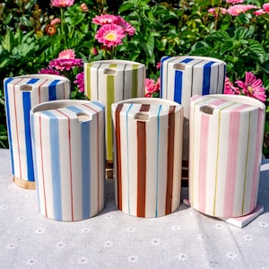 Peut inclure: Six récipients cylindriques en céramique à rayures verticales de différentes couleurs, dont le bleu, le vert, le marron et le rose. Chaque récipient a une ouverture rectangulaire sur le dessus. Ils sont disposés sur une surface blanche à motifs floraux.