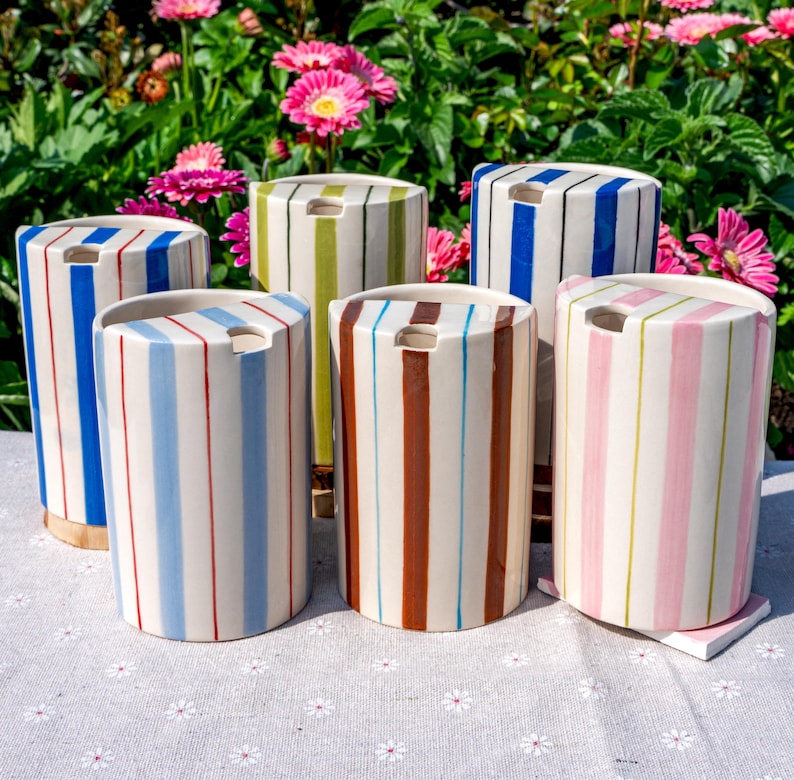May include: A collection of five cylindrical ceramic containers, each featuring vertical stripes in various colors including blue, green, brown, and pink. Each container has a flat top with a small circular opening near the edge for dispensing.