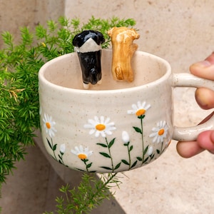 Puede incluir: Una taza de cer&aacute;mica color crema con acabado moteado, que presenta un patr&oacute;n pintado a mano de margaritas blancas con centros amarillos y tallos verdes. Dos peque&ntilde;as figuras de perros esculpidas, una blanca y negra y otra color canela, est&aacute;n posadas en el borde interior.