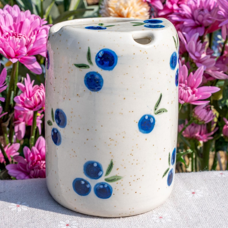 May include: A cylindrical ceramic container with a cream-colored speckled finish. The container is adorned with hand-painted blue blueberries and green leaves. The container has a small opening at the top. The background features pink flowers.