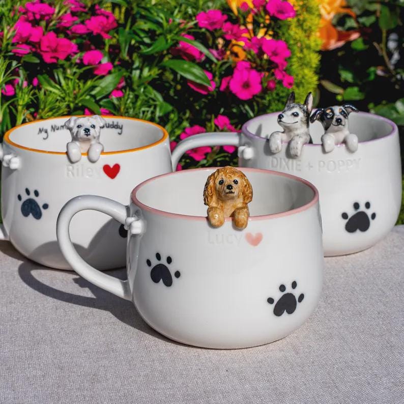 May include: Three white ceramic mugs featuring black paw prints and hand-painted dog figurines perched on the rims. Each mug displays a name and a small heart icon. The mugs have colored rims in orange, pink, and purple, with dog names printed on them.