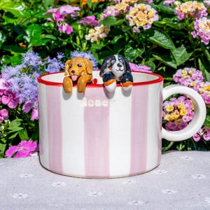May include: A white mug with pink vertical stripes and a red rim. Two ceramic dog figurines are perched on the rim. The word "Money" is printed on the mug. The background features colourful flowers.