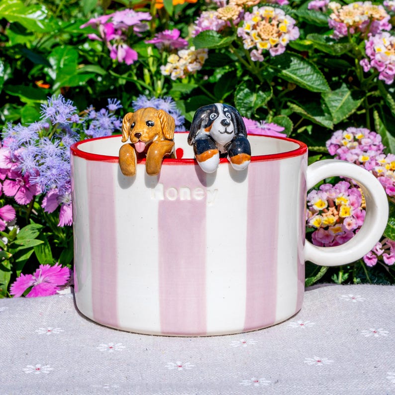 May include: A white mug with pink vertical stripes and a red rim. Two ceramic dog figurines are perched on the rim. The word "Money" is printed on the mug. The background features colourful flowers.
