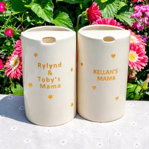 May include: Two off-white ceramic vases with a rectangular shape and a small opening at the top. One vase has the text "Rylnd & Toby's Mama" and the other has "Kellan's Mama" in gold lettering, along with gold star and heart accents.