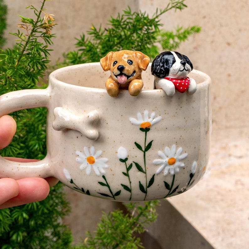 Puede incluir: Una taza de cer&aacute;mica color crema moteada con un detalle de hueso en relieve en el lateral y margaritas blancas pintadas con tallos verdes. Dos peque&ntilde;as figuras de perros esculpidas, una dorada y otra blanca y negra con un pa&ntilde;uelo rojo de lunares, asoman por el borde.
