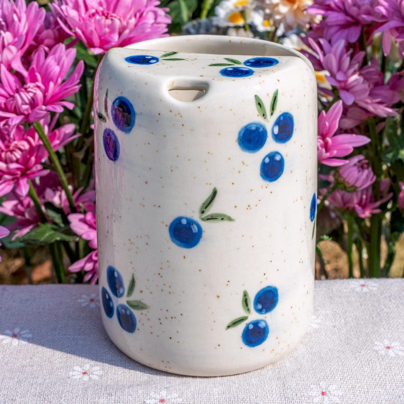 May include: A ceramic toothbrush holder with a cream-colored speckled finish. The holder is decorated with hand-painted blue blueberries and green leaves. It has a rectangular shape with a curved top and a hole for a toothbrush.