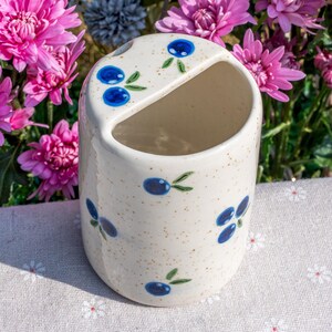 May include: A ceramic toothbrush holder with a speckled, off-white finish. The holder is decorated with blue blueberry designs and green leaves. The holder has a curved opening at the top and a rounded shape. The background features pink flowers.