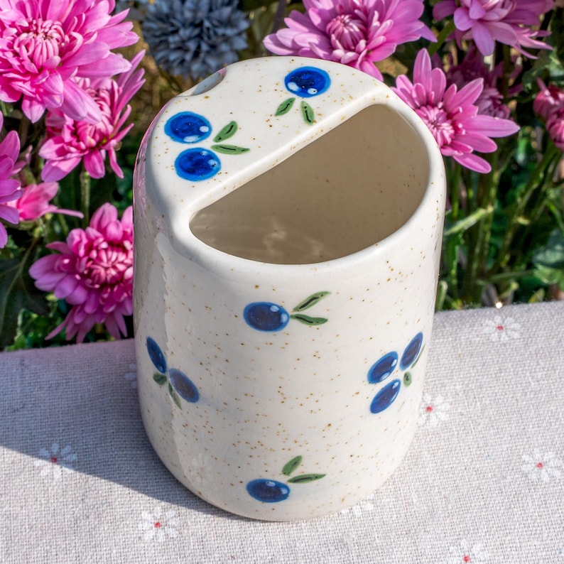 May include: A ceramic toothbrush holder with a speckled, off-white finish. The holder is decorated with blue blueberry designs and green leaves. The holder has a curved opening at the top and a rounded shape. The background features pink flowers.