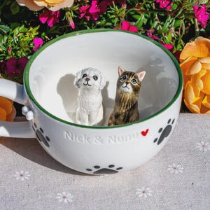 May include: A white ceramic mug featuring a green rim and black paw print patterns on the exterior. Inside the mug are two small figurines: a white dog and a brown tabby cat. The text "Nick & Nunu" with a small red heart is embossed on the front.