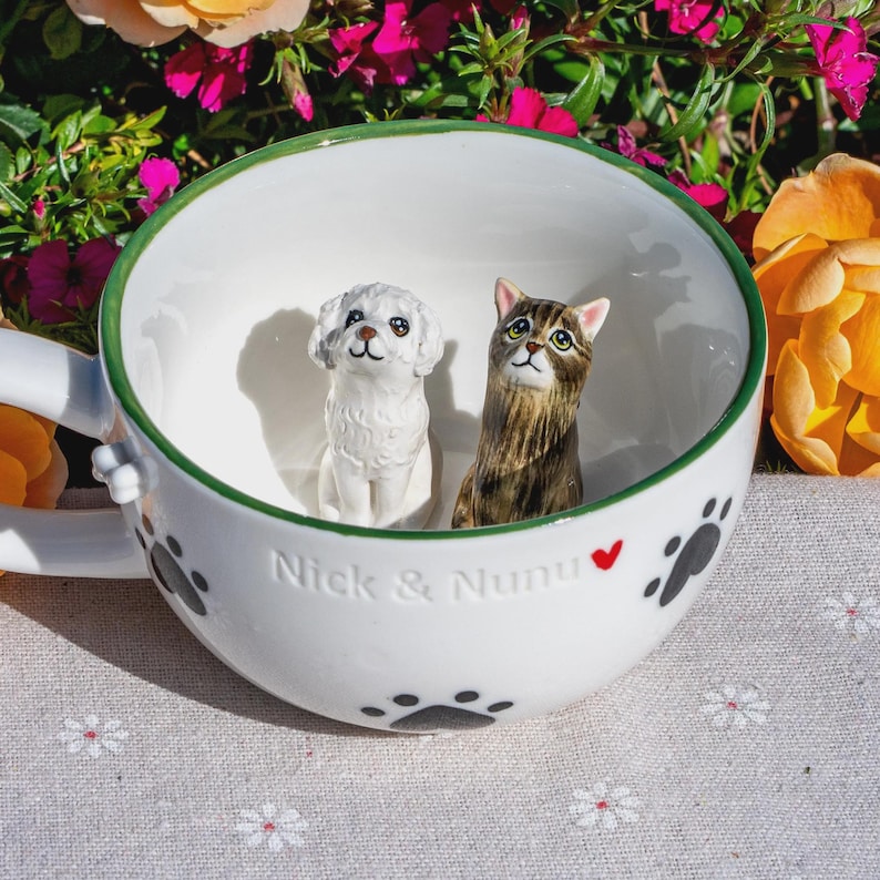 May include: A white ceramic mug featuring a green rim and black paw print patterns on the exterior. Inside the mug are two small figurines: a white dog and a brown tabby cat. The text "Nick & Nunu" with a small red heart is embossed on the front.