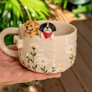 Puede incluir: Una taza de cer&aacute;mica beige con acabado moteado y un patr&oacute;n de margaritas blancas pintadas. Dos peque&ntilde;as figuras de perro esculpidas en tres dimensiones est&aacute;n unidas al borde interior: un perro marr&oacute;n dorado y un perro blanco y negro con un pa&ntilde;uelo rojo.