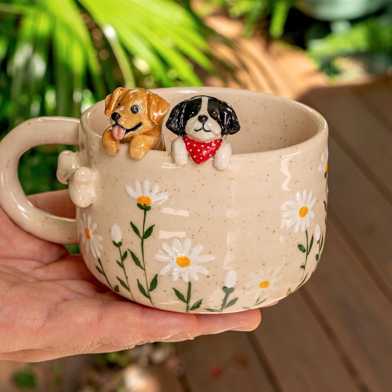 Puede incluir: Una taza de cer&aacute;mica beige con acabado moteado y un patr&oacute;n de margaritas blancas pintadas. Dos peque&ntilde;as figuras de perro esculpidas en tres dimensiones est&aacute;n unidas al borde interior: un perro marr&oacute;n dorado y un perro blanco y negro con un pa&ntilde;uelo rojo.